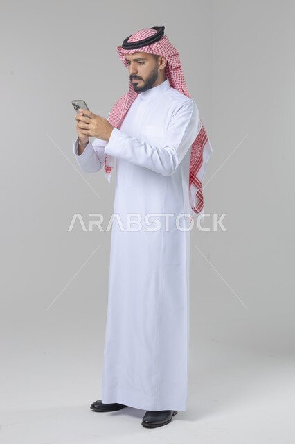  تعابير وايماءات الاندماج والسرور، استخدام التقنية الحديثه المتطورة في الحياة العملية، بورتريه لشاب عربي خليجي سعودي مبتسم يرتدي الشماغ و الثوب التقليدي يقوم بالعمل على الهاتف المحمول، صورة الجسم بالطول الكامل، خلفية بيضاء