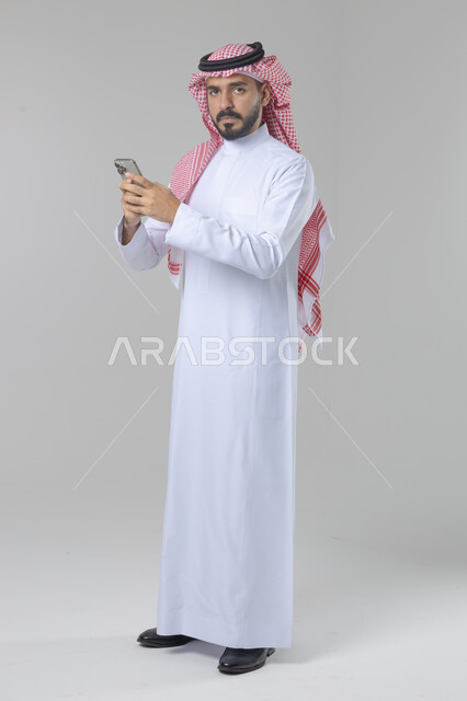  تعابير وايماءات الاندماج والسرور، استخدام التقنية الحديثه المتطورة في الحياة العملية، بورتريه لشاب عربي خليجي سعودي مبتسم يرتدي الشماغ و الثوب التقليدي يقوم بالعمل على الهاتف المحمول، صورة الجسم بالطول الكامل، خلفية بيضاء