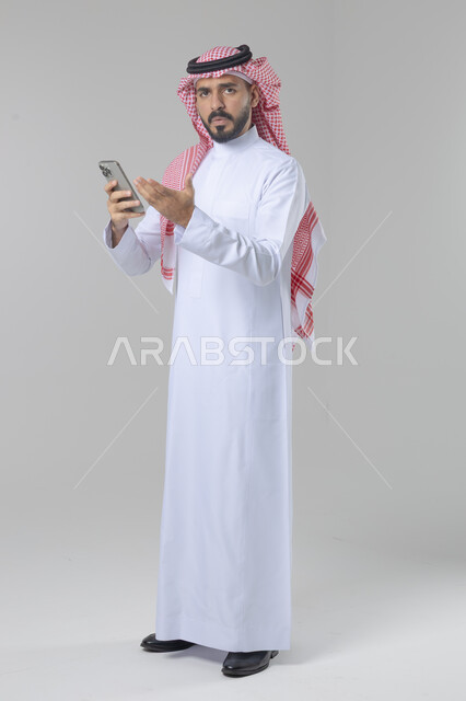  تعابير وايماءات الاندماج والسرور، استخدام التقنية الحديثه المتطورة في الحياة العملية، بورتريه لشاب عربي خليجي سعودي مبتسم يرتدي الشماغ و الثوب التقليدي يقوم بالعمل على الهاتف المحمول، صورة الجسم بالطول الكامل، خلفية بيضاء