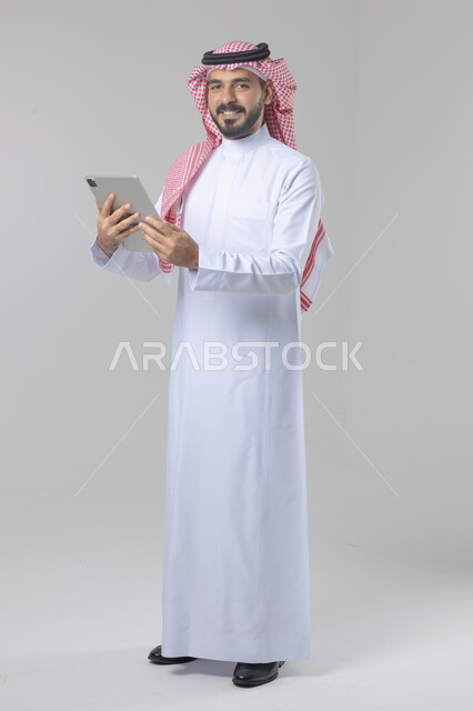 استخدام التقنية الحديثه المتطورة في الحياة العملية عبر الجهاز اللوحي، تعابير وايماءات الاندماج، بورتريه لشاب عربي خليجي سعودي مبتسم يرتدي الشماغ والثوب التقليدي يقوم بالعمل على جهازالتابلت، صورة الجسم بالطول الكامل، خلفية بيضاء