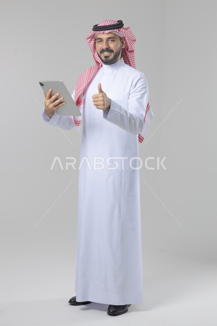 استخدام التقنية الحديثه المتطورة في الحياة العملية عبر الجهاز اللوحي، تعابير وايماءات الاندماج، بورتريه لشاب عربي خليجي سعودي مبتسم يرتدي الشماغ والثوب التقليدي يقوم بالعمل على جهازالتابلت، صورة الجسم بالطول الكامل، خلفية بيضاء