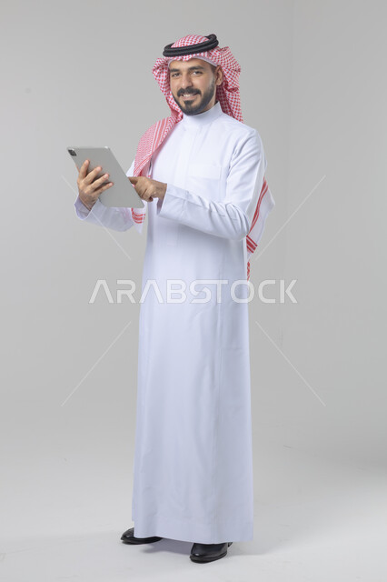 استخدام التقنية الحديثه المتطورة في الحياة العملية عبر الجهاز اللوحي، تعابير وايماءات الاندماج، بورتريه لشاب عربي خليجي سعودي مبتسم يرتدي الشماغ والثوب التقليدي يقوم بالعمل على جهازالتابلت، صورة الجسم بالطول الكامل، خلفية بيضاء