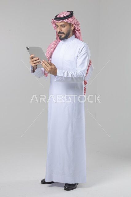 استخدام التقنية الحديثه المتطورة في الحياة العملية عبر الجهاز اللوحي، تعابير وايماءات الاندماج، بورتريه لشاب عربي خليجي سعودي مبتسم يرتدي الشماغ والثوب التقليدي يقوم بالعمل على جهازالتابلت، صورة الجسم بالطول الكامل، خلفية بيضاء
