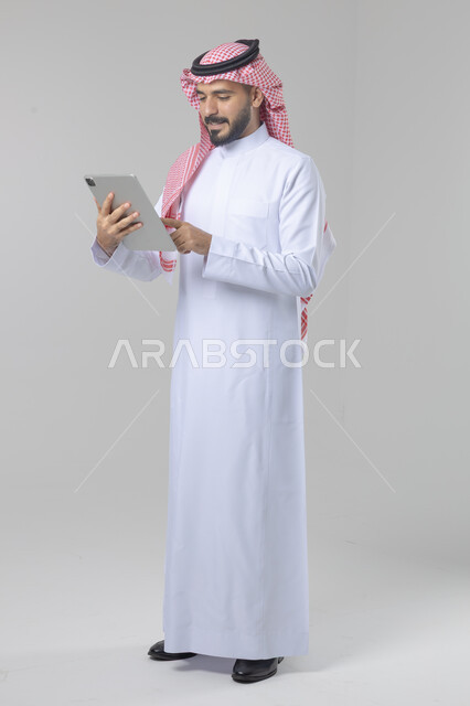 Using modern advanced technology in practical life via tablet, expressions and gestures of integration, portrait of a smiling young Saudi Arabian Gulf man wearing a traditional shemagh and thobe working on a tablet, full body image, white background