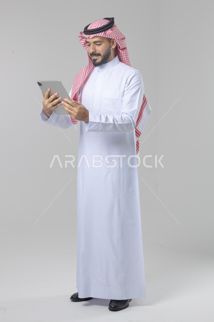 استخدام التقنية الحديثه المتطورة في الحياة العملية عبر الجهاز اللوحي، تعابير وايماءات الاندماج، بورتريه لشاب عربي خليجي سعودي مبتسم يرتدي الشماغ والثوب التقليدي يقوم بالعمل على جهازالتابلت، صورة الجسم بالطول الكامل، خلفية بيضاء