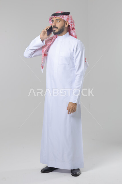 Chatting and conversation using a modern technical device, answering a phone call, portrait of a Saudi Arabian Gulf man wearing traditional thobe and shemagh making a phone call via mobile phone, full body image, white background