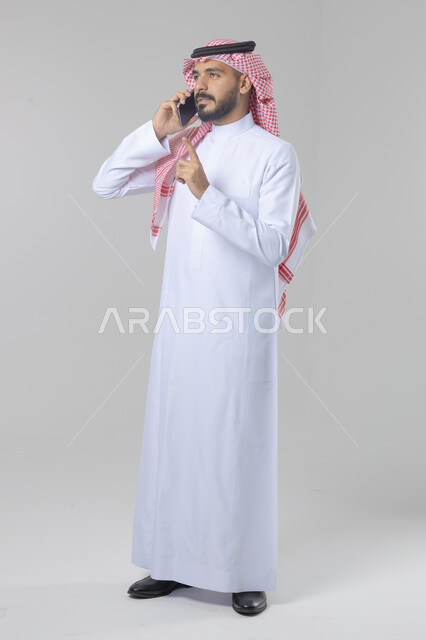 الدردشة والمحادثة باستخدام جهاز تقني حديث, الرد على مكالمة هاتفية، بورتريه لرجل عربي خليجي سعودي يرتدي الثوب التقليدي والشماغ يقوم باجراء اتصال هاتفي من خلال الهاتف المحمول، صورة الجسم بالطول الكامل, خلفية بيضاء