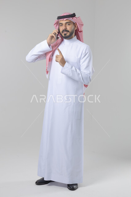 الدردشة والمحادثة باستخدام جهاز تقني حديث, الرد على مكالمة هاتفية، بورتريه لرجل عربي خليجي سعودي يرتدي الثوب التقليدي والشماغ يقوم باجراء اتصال هاتفي من خلال الهاتف المحمول، صورة الجسم بالطول الكامل, خلفية بيضاء