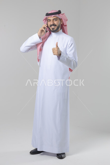 Chatting and conversation using a modern technical device, answering a phone call, portrait of a Saudi Arabian Gulf man wearing traditional thobe and shemagh making a phone call via mobile phone, full body image, white background