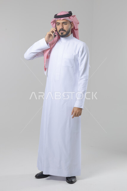 Chatting and conversation using a modern technical device, answering a phone call, portrait of a Saudi Arabian Gulf man wearing traditional thobe and shemagh making a phone call via mobile phone, full body image, white background