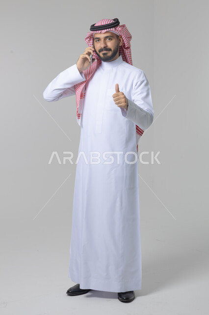 Chatting and conversation using a modern technical device, answering a phone call, portrait of a Saudi Arabian Gulf man wearing traditional thobe and shemagh making a phone call via mobile phone, full body image, white background