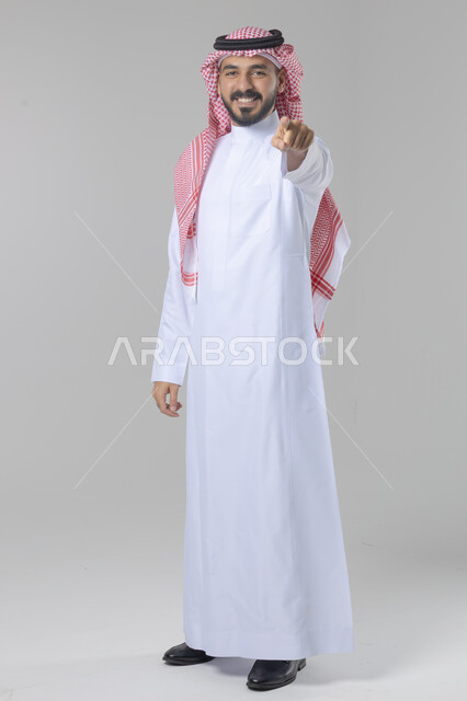 الإشارة إلى شيء ما, الوقوف باستقامة واعتدال, تعابير وايماءات السرور والسعادة، بورتريه لرجل عربي خليجي سعودي يرتدي الثوب التقليدي و الشماغ يشير بيديه لمكان معين, صورة للجسم بالطول الكامل, خلفية بيضاء