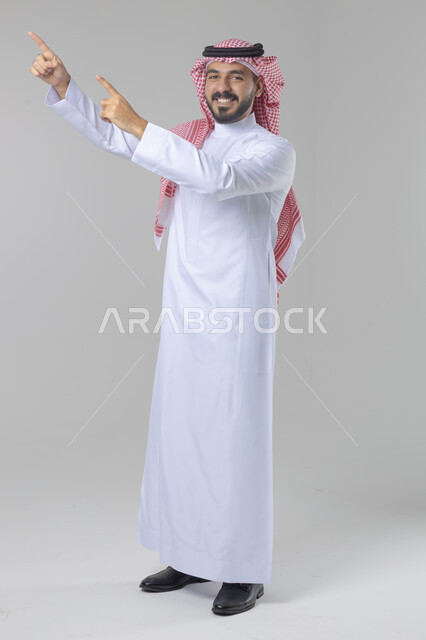 الإشارة إلى شيء ما, الوقوف باستقامة واعتدال, تعابير وايماءات السرور والسعادة، بورتريه لرجل عربي خليجي سعودي يرتدي الثوب التقليدي و الشماغ يشير بيديه لمكان معين, صورة للجسم بالطول الكامل, خلفية بيضاء