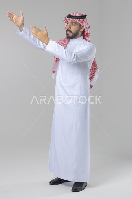 الإشارة إلى شيء ما, الوقوف باستقامة واعتدال, تعابير وايماءات السرور والسعادة، بورتريه لرجل عربي خليجي سعودي يرتدي الثوب التقليدي و الشماغ يشير بيديه لمكان معين, صورة للجسم بالطول الكامل, خلفية بيضاء