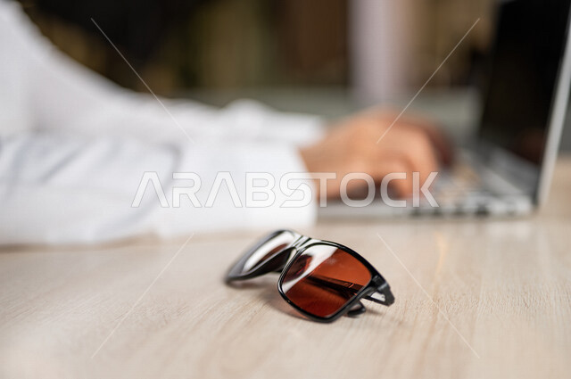 صورة مقربة ليد رجل عربي خليجي سعودي يستخدم الحاسوب المحمول وبجانبه نظارة شمسية، استخدام تقنيات حديثة، انجاز مهام العمل، اعمال مكتبية