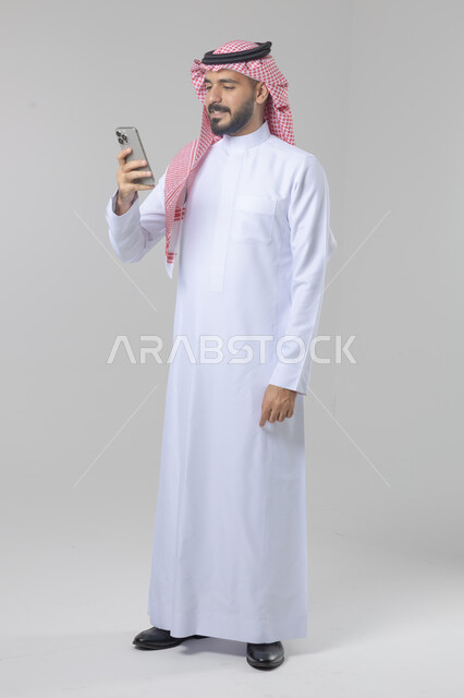  تعابير وايماءات الاندماج والسرور، استخدام التقنية الحديثه المتطورة في الحياة العملية، بورتريه لشاب عربي خليجي سعودي مبتسم يرتدي الشماغ والثوب التقليدي يقوم بالعمل على الهاتف المحمول، صورة الجسم بالطول الكامل، خلفية بيضاء