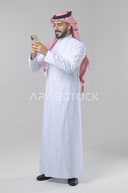  تعابير وايماءات الاندماج والسرور، استخدام التقنية الحديثه المتطورة في الحياة العملية، بورتريه لشاب عربي خليجي سعودي مبتسم يرتدي الشماغ والثوب التقليدي يقوم بالعمل على الهاتف المحمول، صورة الجسم بالطول الكامل، خلفية بيضاء