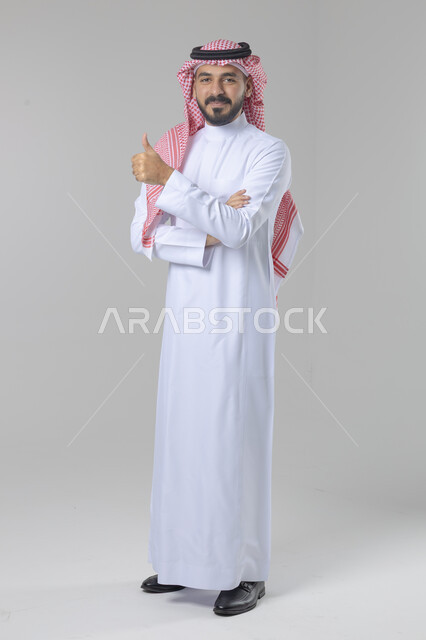  الإمتياز والتميز والنجاح، بورتريه لرجل عربي خليجي سعودي يرتدي الثوب التقليدي والشماغ ويرفع أصابع الإبهام لأعلى، إيماءات السعادة والإعجاب والموافقة، حركة أصبع تدل على القبول والرضا، الجسم بالطول الكامل، خلفية بيضاء 