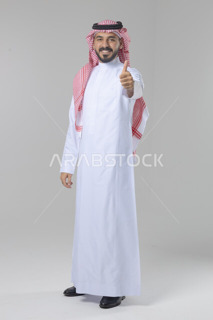  الإمتياز والتميز والنجاح، بورتريه لرجل عربي خليجي سعودي يرتدي الثوب التقليدي والشماغ ويرفع أصابع الإبهام لأعلى، إيماءات السعادة والإعجاب والموافقة، حركة أصبع تدل على القبول والرضا، الجسم بالطول الكامل، خلفية بيضاء 