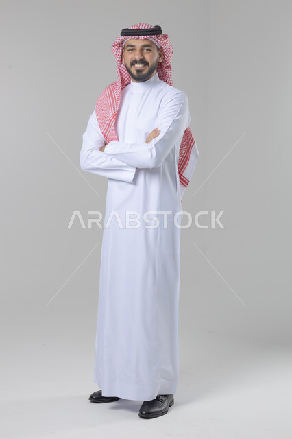 الاعتناء والاهتمام بالمظهر الخارجي، مفهوم الاناقة والرجولة، التقاط صور شخصية للوثائق الرسمية، بورتريه لشاب عربي خليجي سعودي يرتدي الثوب التقليدي والشماغ يقف بأيدي متقاطعة ينظر الى الكاميرا بتعابير السرور والثقة بالنفس، الجسم بالطول الكامل، خلفية بيضاء