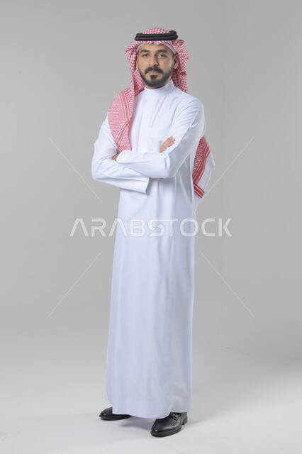 الاعتناء والاهتمام بالمظهر الخارجي، مفهوم الاناقة والرجولة، التقاط صور شخصية للوثائق الرسمية، بورتريه لشاب عربي خليجي سعودي يرتدي الثوب التقليدي والشماغ يقف بأيدي متقاطعة ينظر الى الكاميرا بتعابير السرور والثقة بالنفس، الجسم بالطول الكامل، خلفية بيضاء