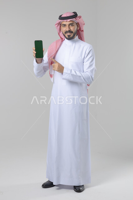 عرض شاشة خضراء كروما فارغة على الموبايل، بورتريه لرجل عربي خليجي سعودي يرتدي الشماغ والثوب التقليدي يمسك بيده الهاتف المحمول، دمج التقنية والتكنولوجيا بالحياة اليومية، صورة الجسم بالطول الكامل، خلفية بيضاء