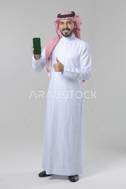 عرض شاشة خضراء كروما فارغة على الموبايل، بورتريه لرجل عربي خليجي سعودي يرتدي الشماغ والثوب التقليدي يمسك بيده الهاتف المحمول، دمج التقنية والتكنولوجيا بالحياة اليومية، صورة الجسم بالطول الكامل، خلفية بيضاء