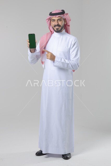 عرض شاشة خضراء كروما فارغة على الموبايل، بورتريه لرجل عربي خليجي سعودي يرتدي الشماغ والثوب التقليدي يمسك بيده الهاتف المحمول، دمج التقنية والتكنولوجيا بالحياة اليومية، صورة الجسم بالطول الكامل، خلفية بيضاء