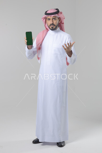 عرض شاشة خضراء كروما فارغة على الموبايل، بورتريه لرجل عربي خليجي سعودي يرتدي الشماغ والثوب التقليدي يمسك بيده الهاتف المحمول، دمج التقنية والتكنولوجيا بالحياة اليومية، صورة الجسم بالطول الكامل، خلفية بيضاء