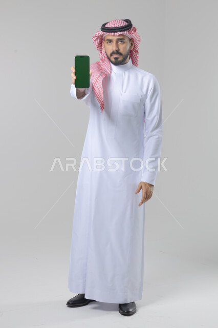 عرض شاشة خضراء كروما فارغة على الموبايل، بورتريه لرجل عربي خليجي سعودي يرتدي الشماغ والثوب التقليدي يمسك بيده الهاتف المحمول، دمج التقنية والتكنولوجيا بالحياة اليومية، صورة الجسم بالطول الكامل، خلفية بيضاء