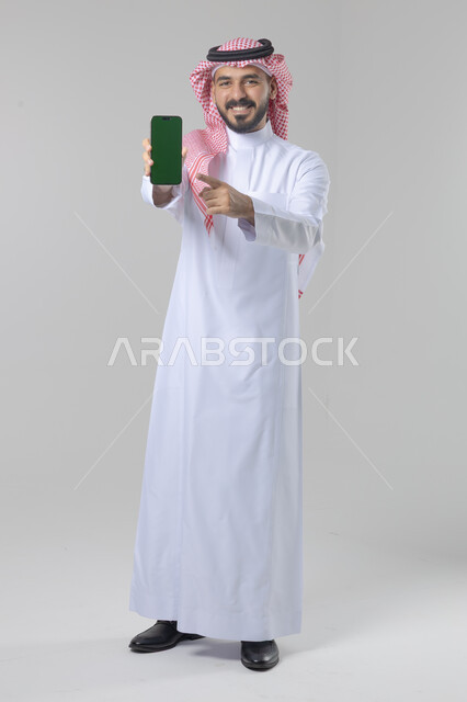 عرض شاشة خضراء كروما فارغة على الموبايل، بورتريه لرجل عربي خليجي سعودي يرتدي الشماغ والثوب التقليدي يمسك بيده الهاتف المحمول، دمج التقنية والتكنولوجيا بالحياة اليومية، صورة الجسم بالطول الكامل، خلفية بيضاء