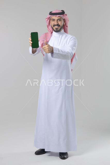 عرض شاشة خضراء كروما فارغة على الموبايل، بورتريه لرجل عربي خليجي سعودي يرتدي الشماغ والثوب التقليدي يمسك بيده الهاتف المحمول، دمج التقنية والتكنولوجيا بالحياة اليومية، صورة الجسم بالطول الكامل، خلفية بيضاء