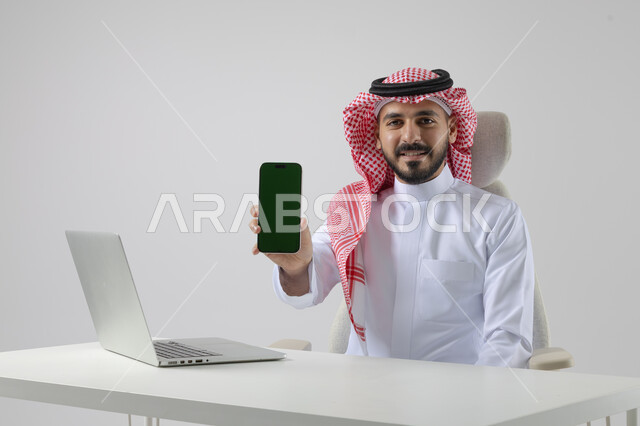 عرض شاشة خضراء كروما فارغة على الهاتف المحمول، بورتريه مقرب لرجل عربي خليجي سعودي يرتدي الشماغ والثوب التقليدي يعمل على الحاسب المحمول، دمج التقنية والتكنولوجيا بالحياة العملية، خلفية بيضاء