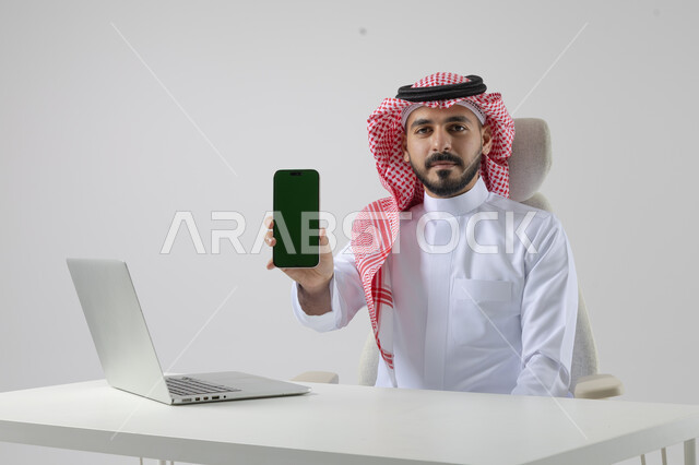 عرض شاشة خضراء كروما فارغة على الهاتف المحمول، بورتريه مقرب لرجل عربي خليجي سعودي يرتدي الشماغ والثوب التقليدي يعمل على الحاسب المحمول، دمج التقنية والتكنولوجيا بالحياة العملية، خلفية بيضاء