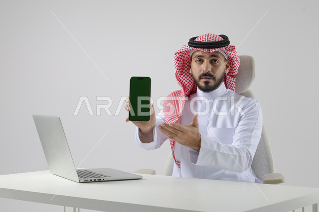 عرض شاشة خضراء كروما فارغة على الهاتف المحمول، بورتريه مقرب لرجل عربي خليجي سعودي يرتدي الشماغ والثوب التقليدي يعمل على الحاسب المحمول، دمج التقنية والتكنولوجيا بالحياة العملية، خلفية بيضاء