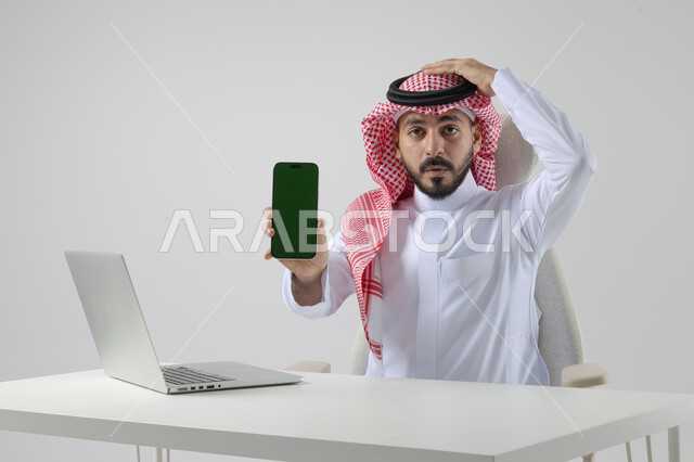 Blank green screen chroma key on mobile phone, close-up portrait of Saudi Arabian Gulf man wearing traditional keffiyeh and thobe working on laptop, technology integration with business life, white background