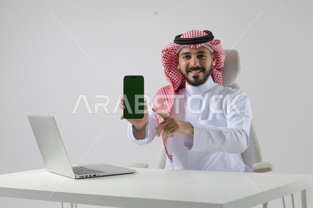 عرض شاشة خضراء كروما فارغة على الهاتف المحمول، بورتريه مقرب لرجل عربي خليجي سعودي يرتدي الشماغ والثوب التقليدي يعمل على الحاسب المحمول، دمج التقنية والتكنولوجيا بالحياة العملية، خلفية بيضاء