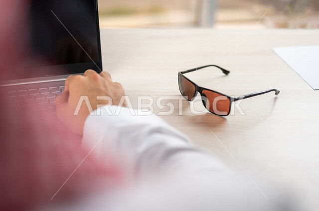 Close-up of the hand of a Saudi Arabian Gulf man using a laptop computer with sunglasses next to him, using modern technologies, completing work tasks, office work