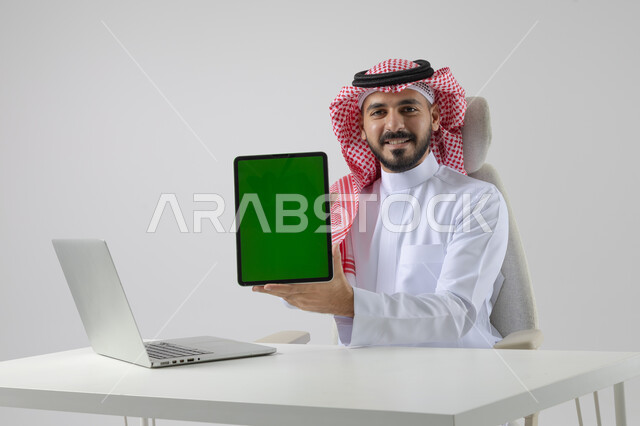 عرض شاشة خضراء كروما فارغة على جهاز التابلت، بورتريه مقرب لرجل عربي خليجي سعودي يرتدي الشماغ والثوب التقليدي يعمل على الحاسب المحمول، دمج التقنية والتكنولوجيا بالحياة العملية، خلفية بيضاء