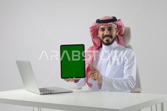عرض شاشة خضراء كروما فارغة على جهاز التابلت، بورتريه مقرب لرجل عربي خليجي سعودي يرتدي الشماغ والثوب التقليدي يعمل على الحاسب المحمول، دمج التقنية والتكنولوجيا بالحياة العملية، خلفية بيضاء