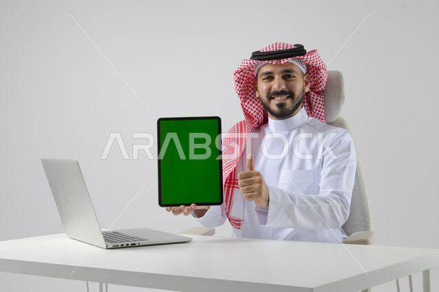 Blank green screen chroma key display on tablet, close-up portrait of Saudi Arabian Gulf man wearing traditional keffiyeh and thobe working on laptop, technology integration with business life, white background