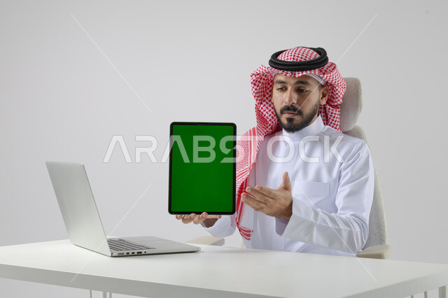 عرض شاشة خضراء كروما فارغة على جهاز التابلت، بورتريه مقرب لرجل عربي خليجي سعودي يرتدي الشماغ والثوب التقليدي يعمل على الحاسب المحمول، دمج التقنية والتكنولوجيا بالحياة العملية، خلفية بيضاء
