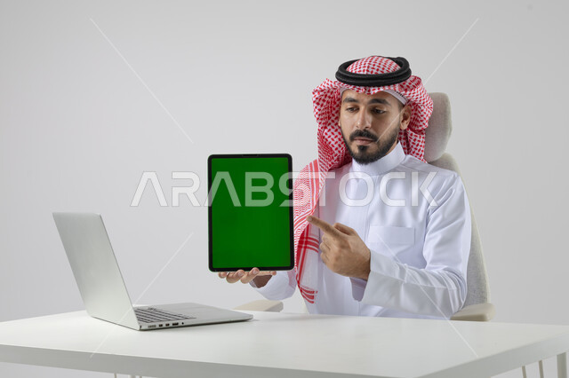 عرض شاشة خضراء كروما فارغة على جهاز التابلت، بورتريه مقرب لرجل عربي خليجي سعودي يرتدي الشماغ والثوب التقليدي يعمل على الحاسب المحمول، دمج التقنية والتكنولوجيا بالحياة العملية، خلفية بيضاء