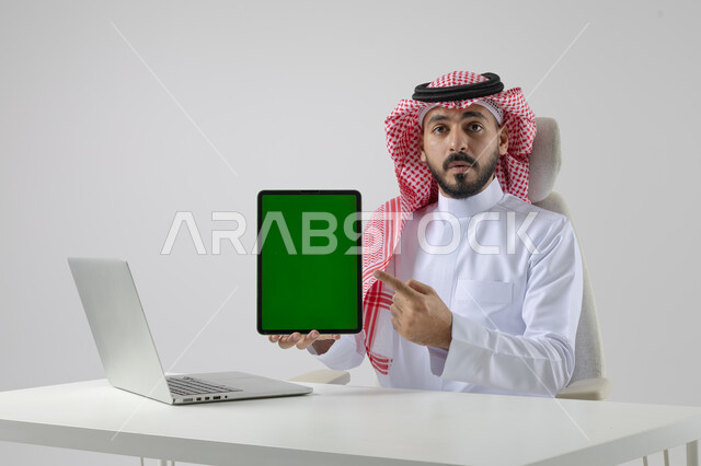 عرض شاشة خضراء كروما فارغة على جهاز التابلت، بورتريه مقرب لرجل عربي خليجي سعودي يرتدي الشماغ والثوب التقليدي يعمل على الحاسب المحمول، دمج التقنية والتكنولوجيا بالحياة العملية، خلفية بيضاء