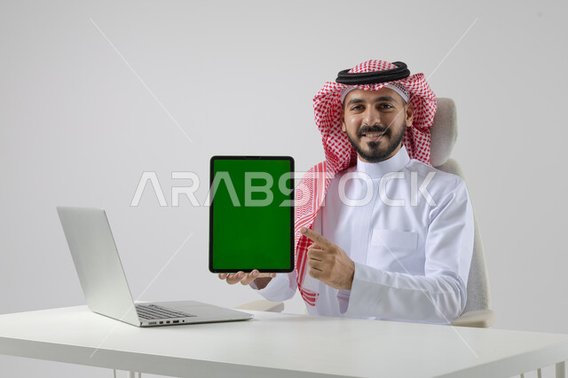 عرض شاشة خضراء كروما فارغة على جهاز التابلت، بورتريه مقرب لرجل عربي خليجي سعودي يرتدي الشماغ والثوب التقليدي يعمل على الحاسب المحمول، دمج التقنية والتكنولوجيا بالحياة العملية، خلفية بيضاء