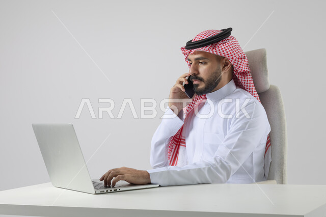 الرد والاجابة على الاتصالات الهاتفية عبر الهاتف المحمول، بورتريه مقرب لرجل عربي خليجي سعودي يرتدي الشماغ والثوب التقليدي يعمل على الحاسب المحمول، دمج التقنية والتكنولوجيا بالحياة العملية، خلفية بيضاء