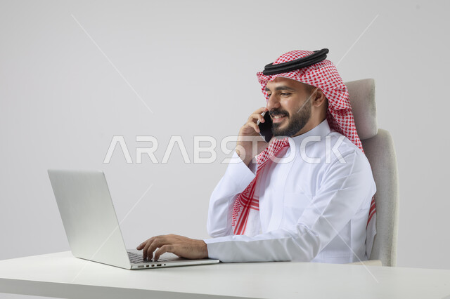 الرد والاجابة على الاتصالات الهاتفية عبر الهاتف المحمول، بورتريه مقرب لرجل عربي خليجي سعودي يرتدي الشماغ والثوب التقليدي يعمل على الحاسب المحمول، دمج التقنية والتكنولوجيا بالحياة العملية، خلفية بيضاء