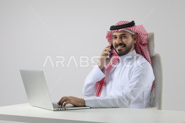 الرد والاجابة على الاتصالات الهاتفية عبر الهاتف المحمول، بورتريه مقرب لرجل عربي خليجي سعودي يرتدي الشماغ والثوب التقليدي يعمل على الحاسب المحمول، دمج التقنية والتكنولوجيا بالحياة العملية، خلفية بيضاء