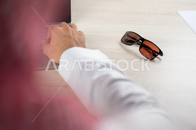 صورة مقربة ليد رجل عربي خليجي سعودي يستخدم الحاسوب المحمول وبجانبه نظارة شمسية، استخدام تقنيات حديثة، انجاز مهام العمل، اعمال مكتبية