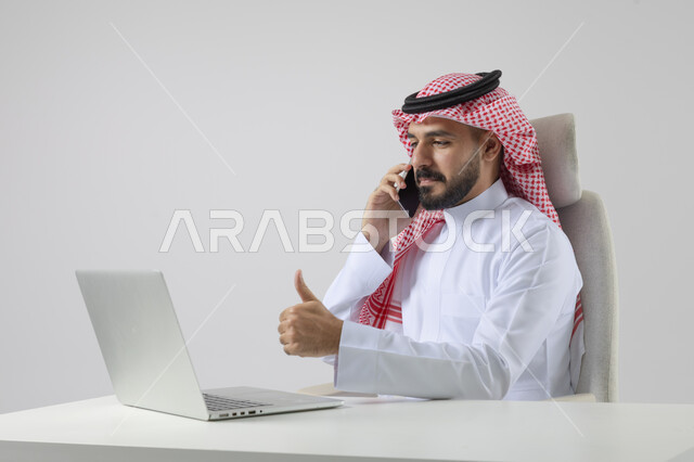 الرد والاجابة على الاتصالات الهاتفية عبر الهاتف المحمول، بورتريه مقرب لرجل عربي خليجي سعودي يرتدي الشماغ والثوب التقليدي يعمل على الحاسب المحمول، دمج التقنية والتكنولوجيا بالحياة العملية، خلفية بيضاء